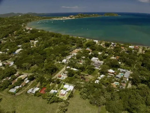 Vista aérea de gran altitud de una zona costera con árboles verdes, edificios y una bahía con botes bajo un cielo azul.