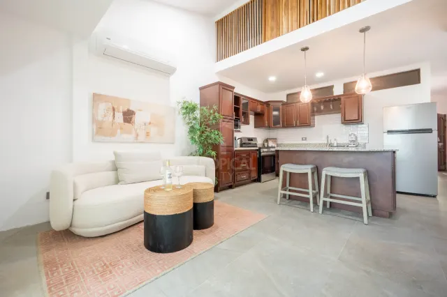 Sala de concepto abierto con un sofá crema, una alfombra rosada y dos mesas auxiliares redondas. La cocina integrada tiene gabinetes de madera oscura y una barra con dos taburetes.