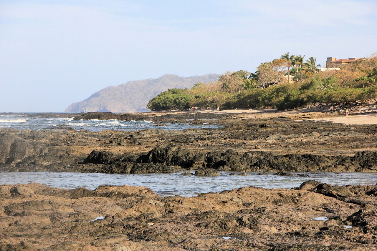Plataforma rocosa y arena clara en Playa Langosta, Guanacaste, con casas frente al mar entre las palmeras